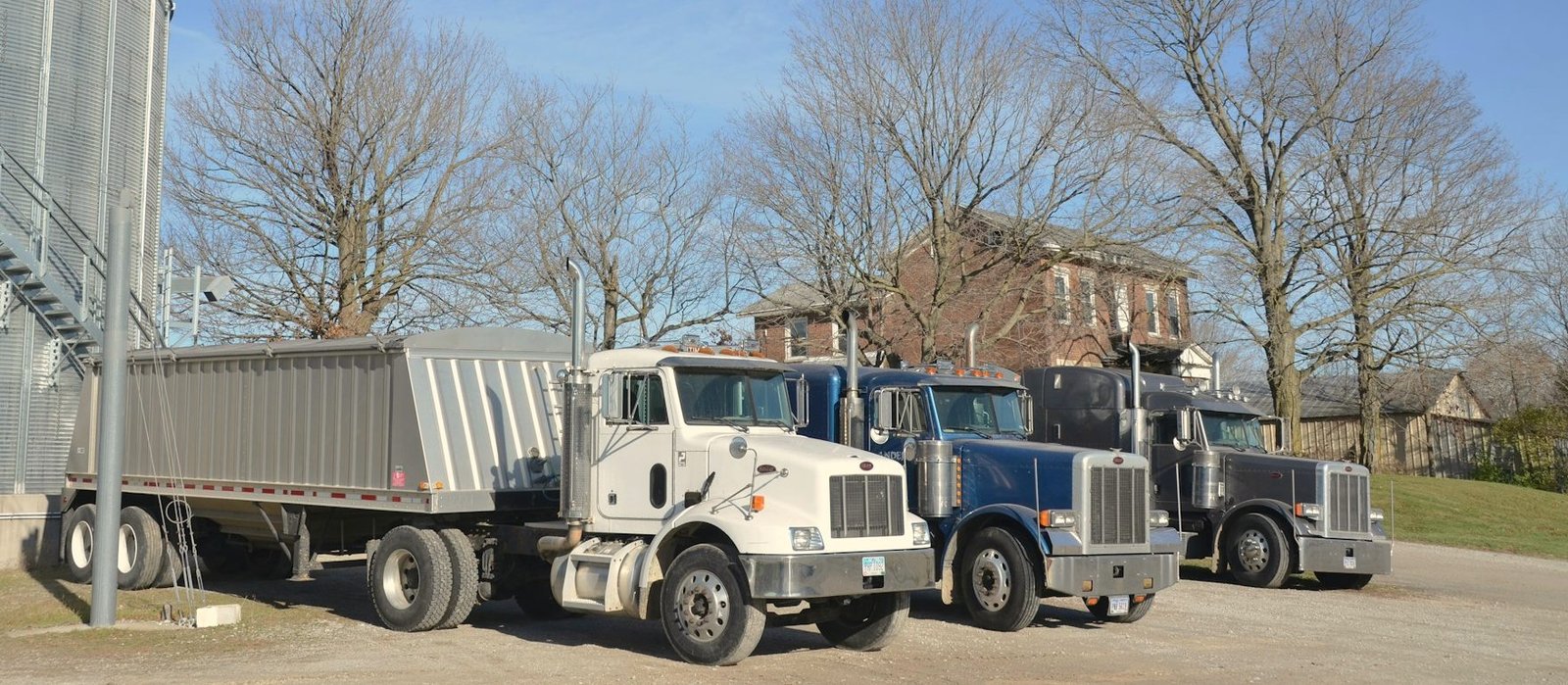 Three semi tractors with a grain hopper trailer parked beside a steel grain bin